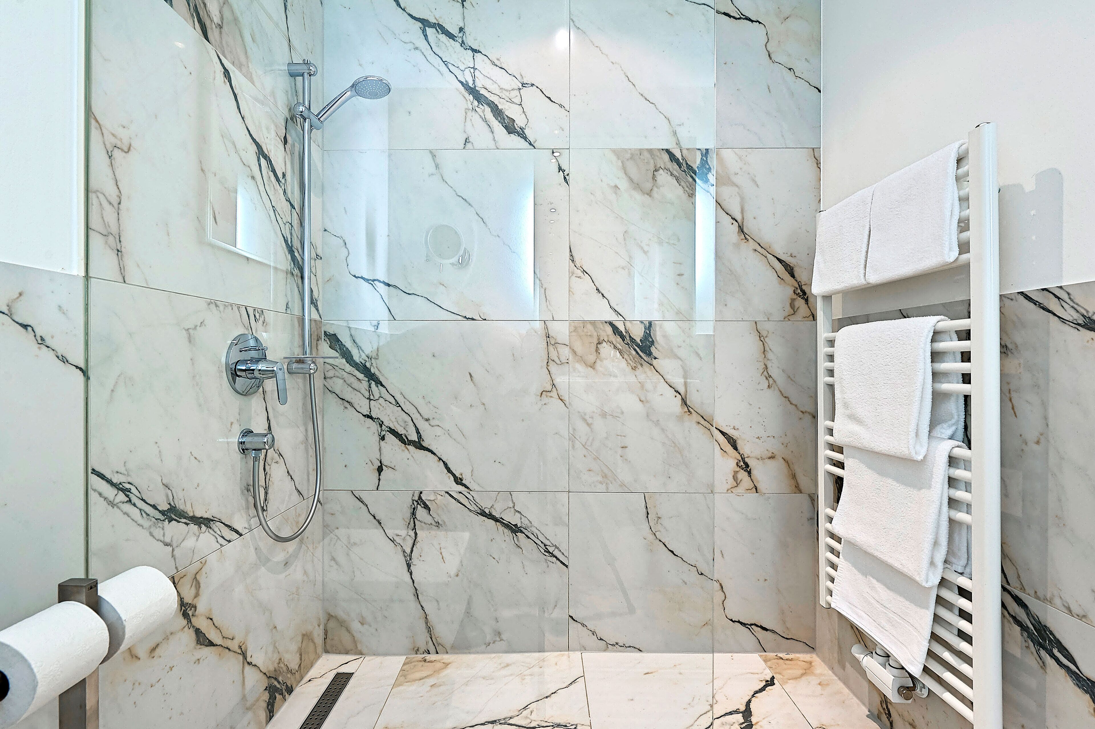 A bathroom with a glass shower enclosure, marble walls, a towel rack, and a toilet paper holder.