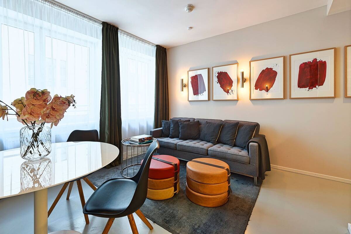 A modern living room with a round dining table, black chairs, a grey sofa, and orange ottomans. There are framed abstract paintings on the wall and a vase with flowers on the table.