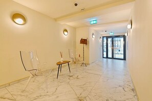 A hotel lobby with marble flooring, clear chairs, a small table with a vase, and a coat rack.