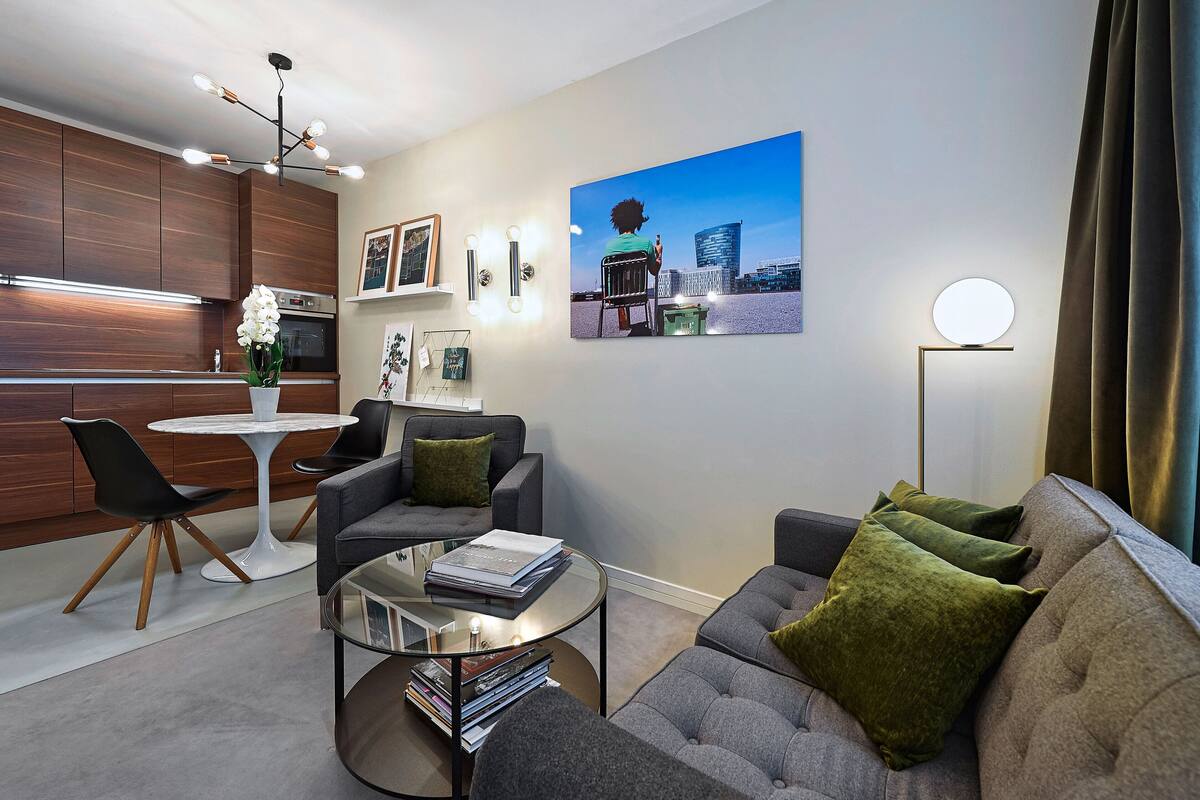 A modern living room with a grey sofa, a coffee table, and a dining area with a white table and chairs. There is a painting on the wall and a floor lamp.