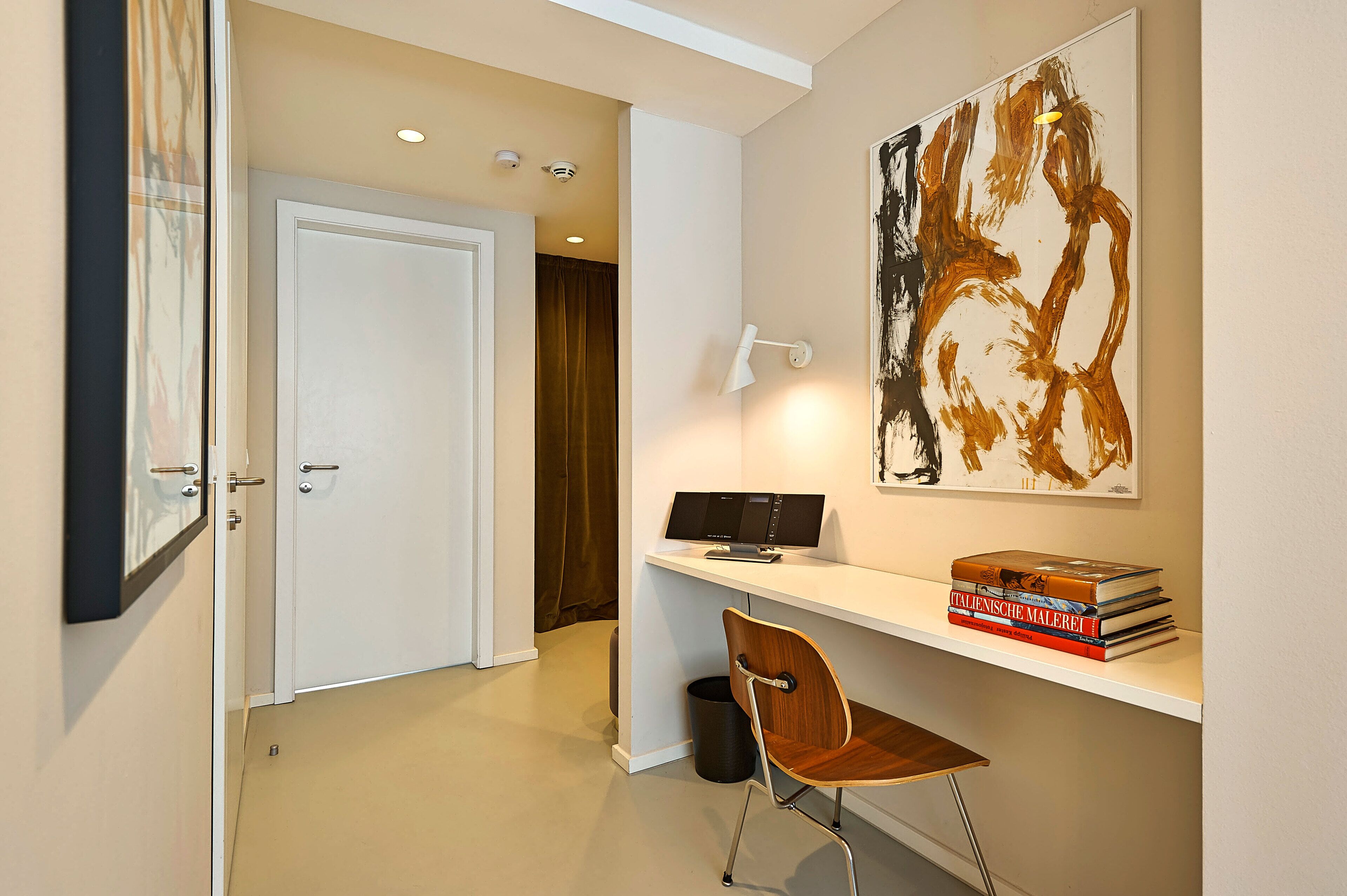 A modern room with a white desk, a chair, a bookshelf with books, a painting, and a door leading to another room.