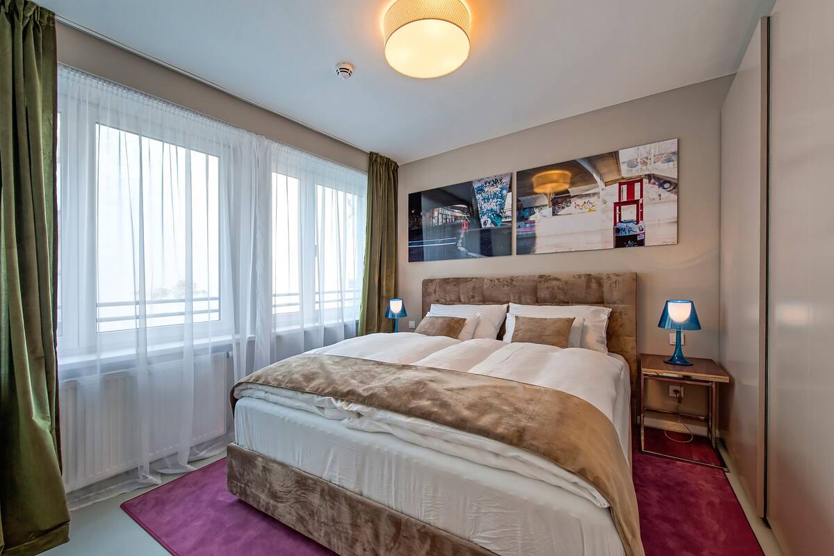 A well-lit bedroom with a large bed, a nightstand with a lamp, and artwork on the wall.
