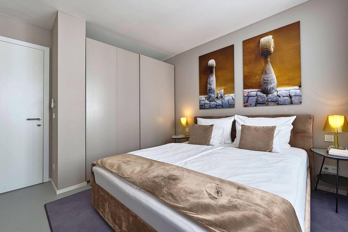 A modern hotel room with a large bed, two bedside tables with lamps, a wardrobe, and two paintings on the wall.