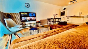A modern living room with a sofa, a TV displaying a movie, a clock, and a kitchen area. - Nivas - 2 rooms, kitchen, bathroom, 6 persons (Montabaur)