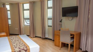 A hotel room with a bed, a desk, a chair, a TV, and a window with curtains. - Tropical Plaza Apartments (Kigali)