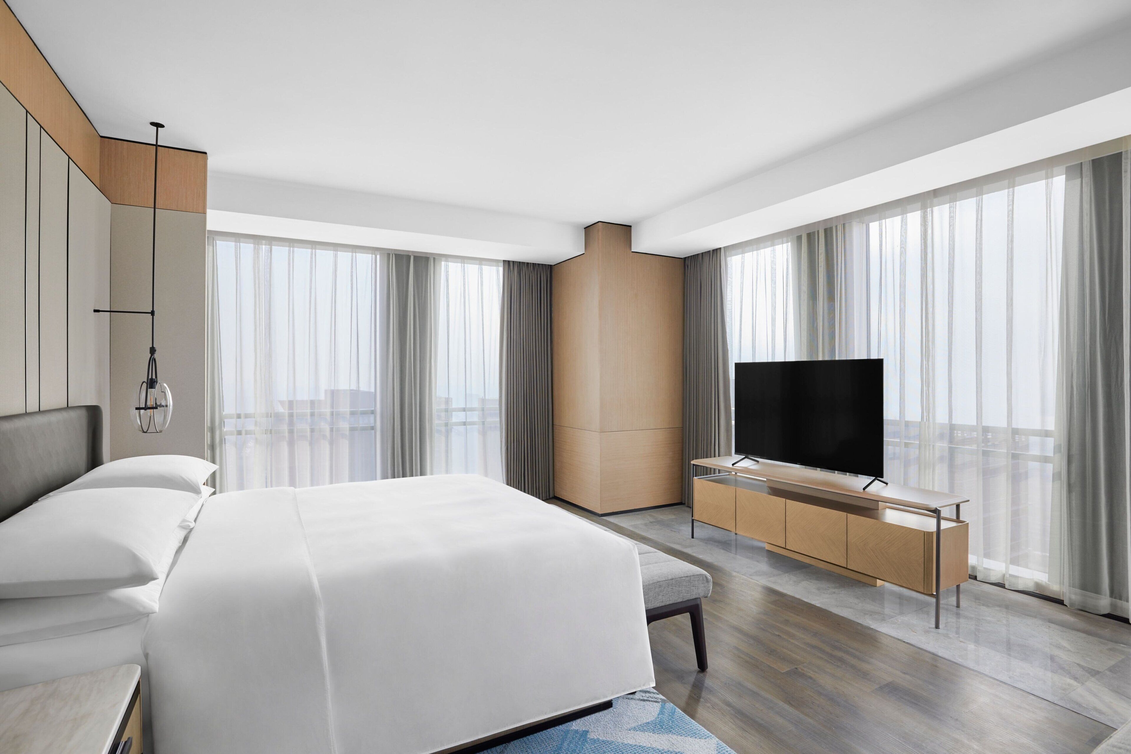 Een moderne hotelkamer met een groot bed, een flatscreen televisie, een houten kast en grote ramen met dunne gordijnen.