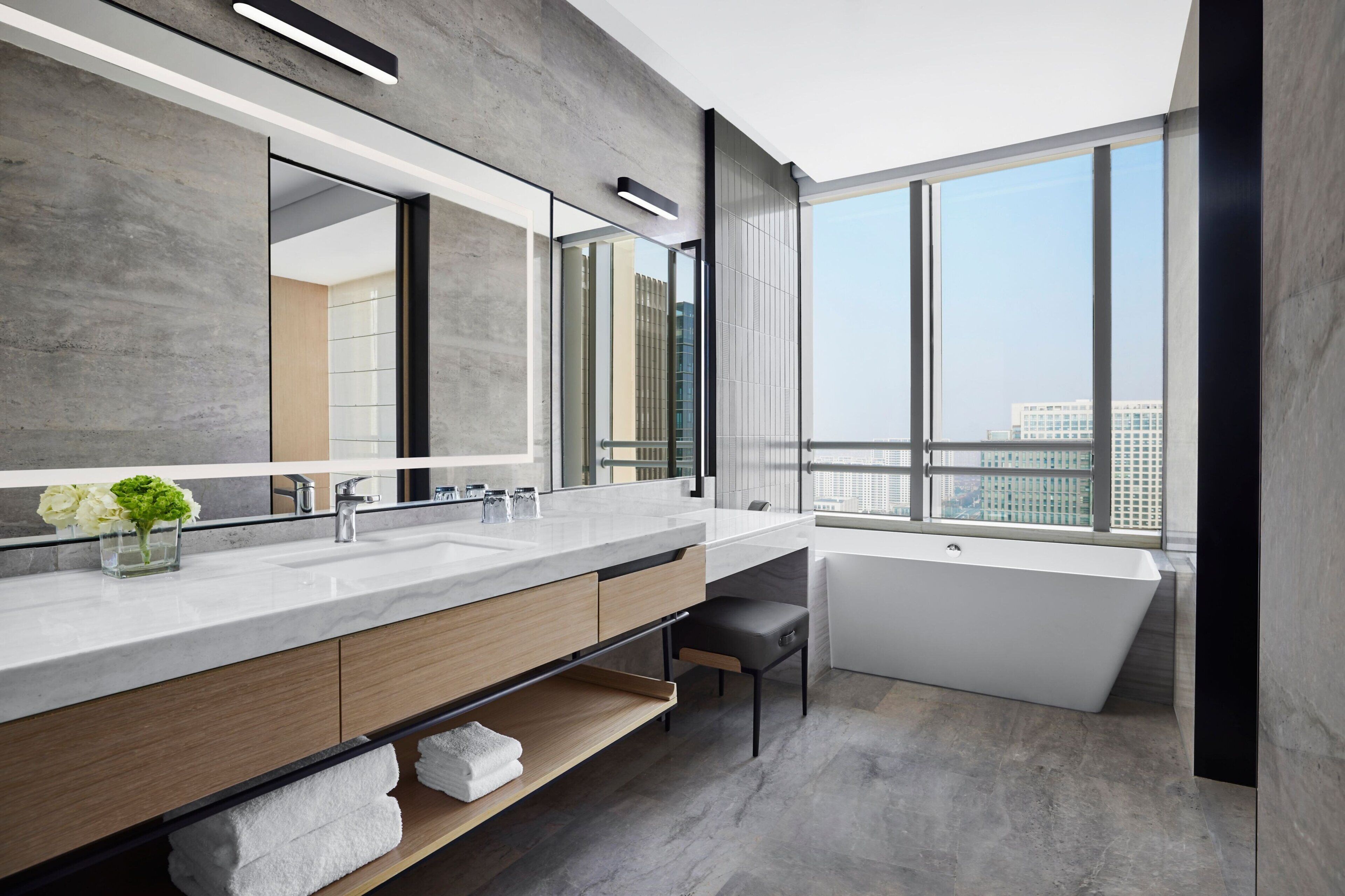Un baño moderno con un espejo grande, dos lavamanos, una bañera y vistas de la ciudad a través de amplias ventanas.