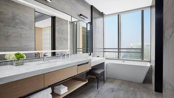 Un baño moderno con un espejo grande, dos lavamanos, una bañera y vistas de la ciudad a través de amplias ventanas.
