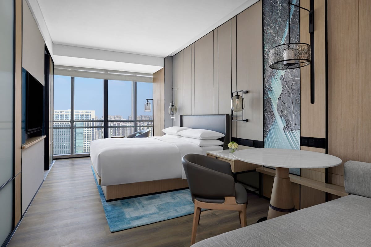 Une chambre d’hôtel moderne dotée d’un grand lit, d’un bureau, d’une chaise et offrant une vue sur la ville grâce à de grandes fenêtres.