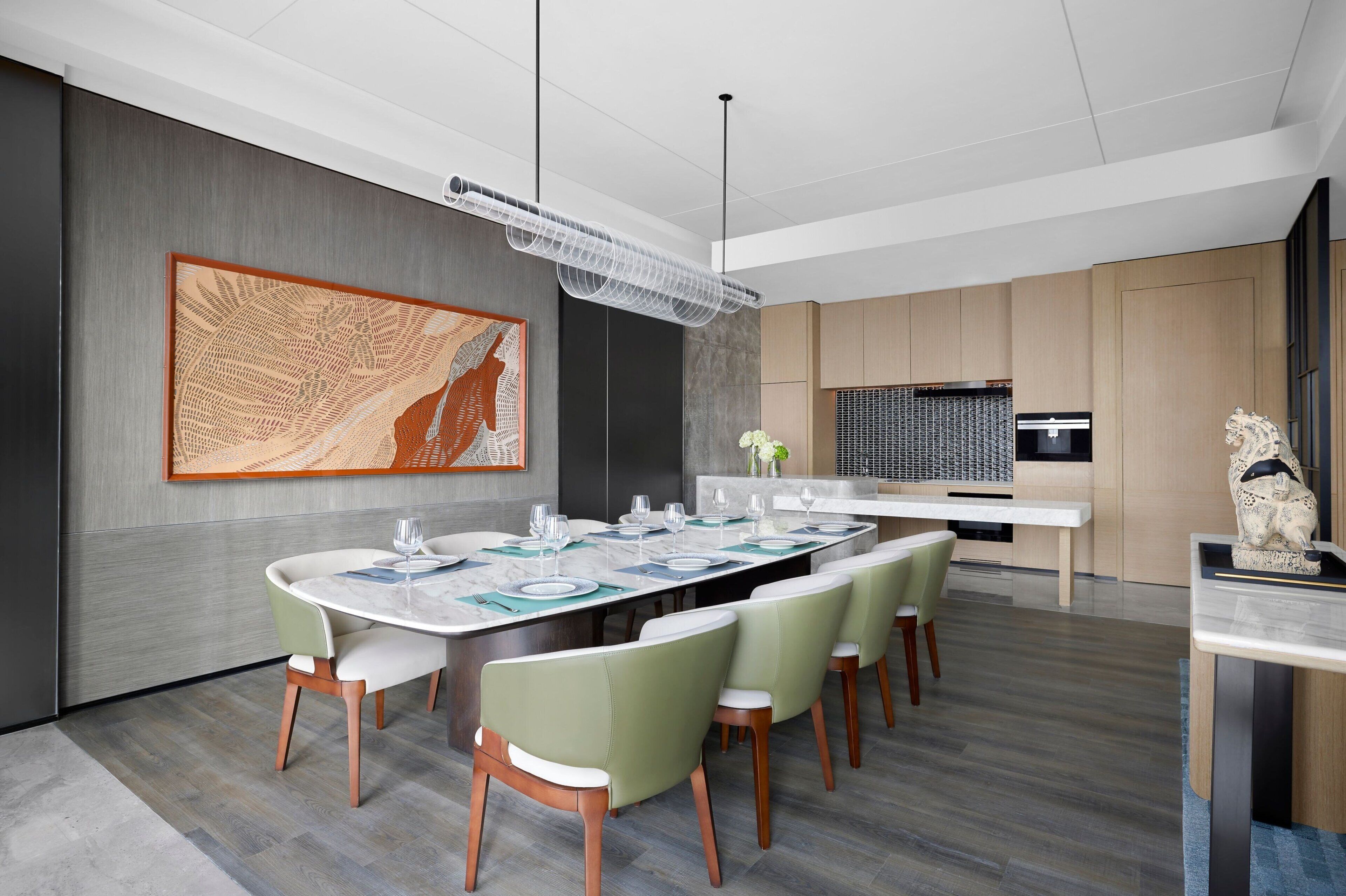 Un comedor moderno con una mesa grande dispuesta para seis personas, rodeada de sillas verdes, un cuadro abstracto de gran tamaño en la pared y una cocina al fondo.