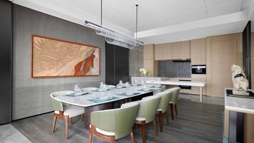 Un comedor moderno con una mesa grande dispuesta para seis personas, rodeada de sillas verdes, un cuadro abstracto de gran tamaño en la pared y una cocina al fondo.