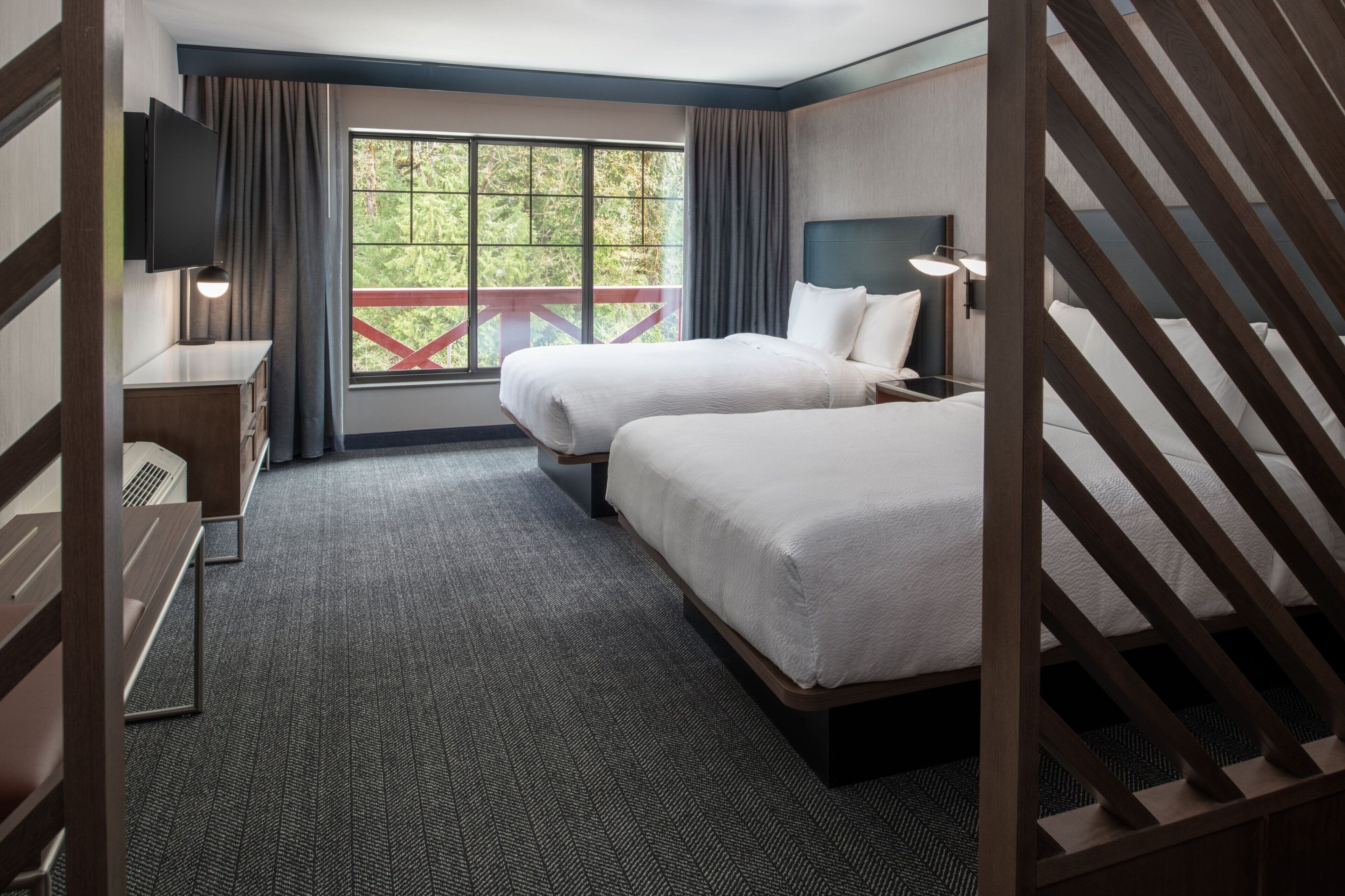 A hotel room with two beds, a desk, a TV, and a window with a view of greenery.