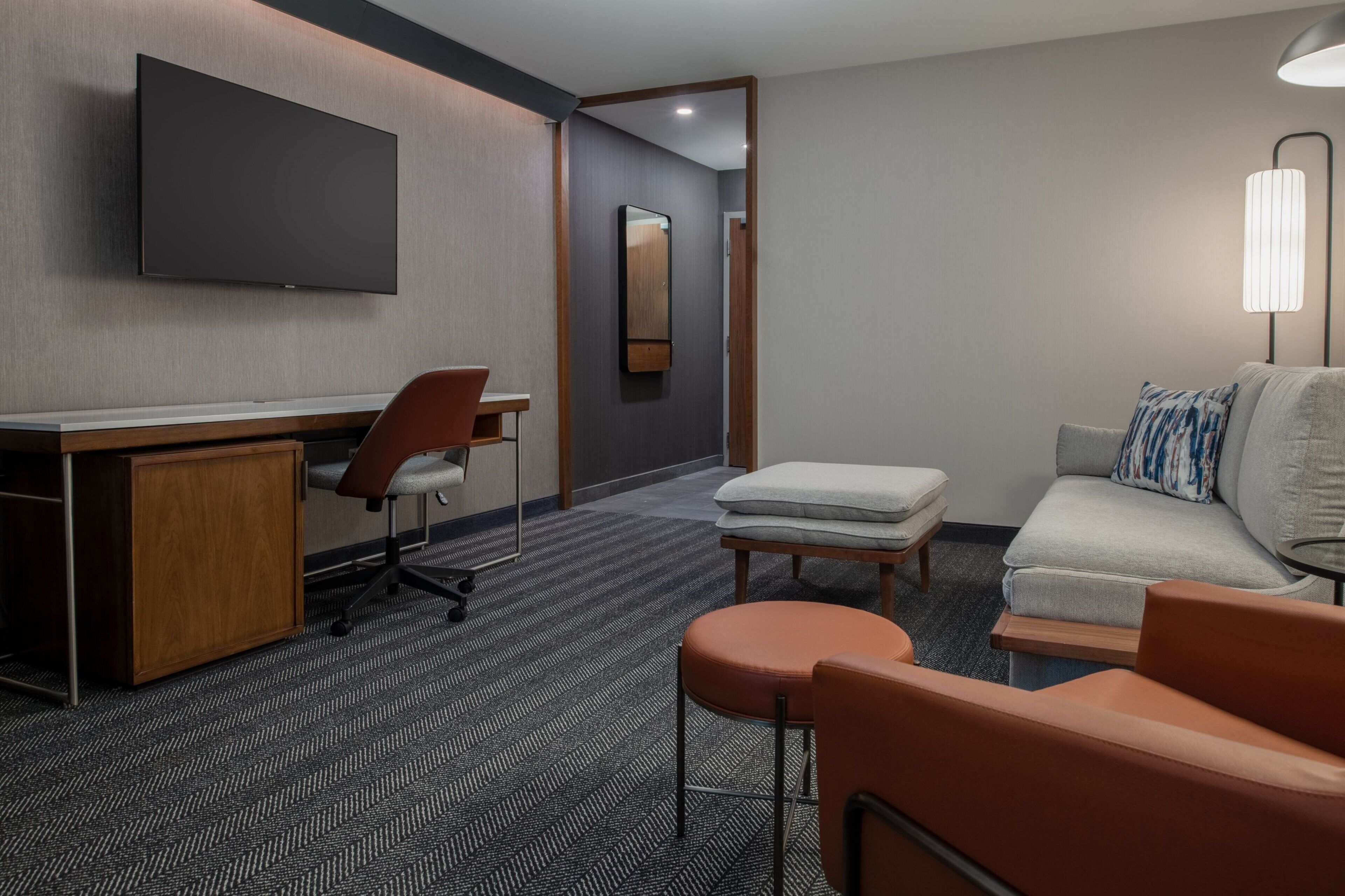 A hotel room with a flat-screen TV mounted on the wall, a desk with a chair, a sofa with a pillow, a small table, and a lamp.
