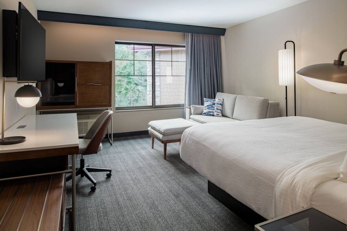 Een hotelkamer met een bed, een bureau met stoel, een televisie, een raam met gordijnen en een lamp.