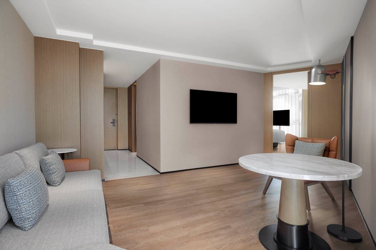 A modern living room with a round marble table, a sofa with cushions, a flat-screen TV mounted on the wall, and a floor lamp.