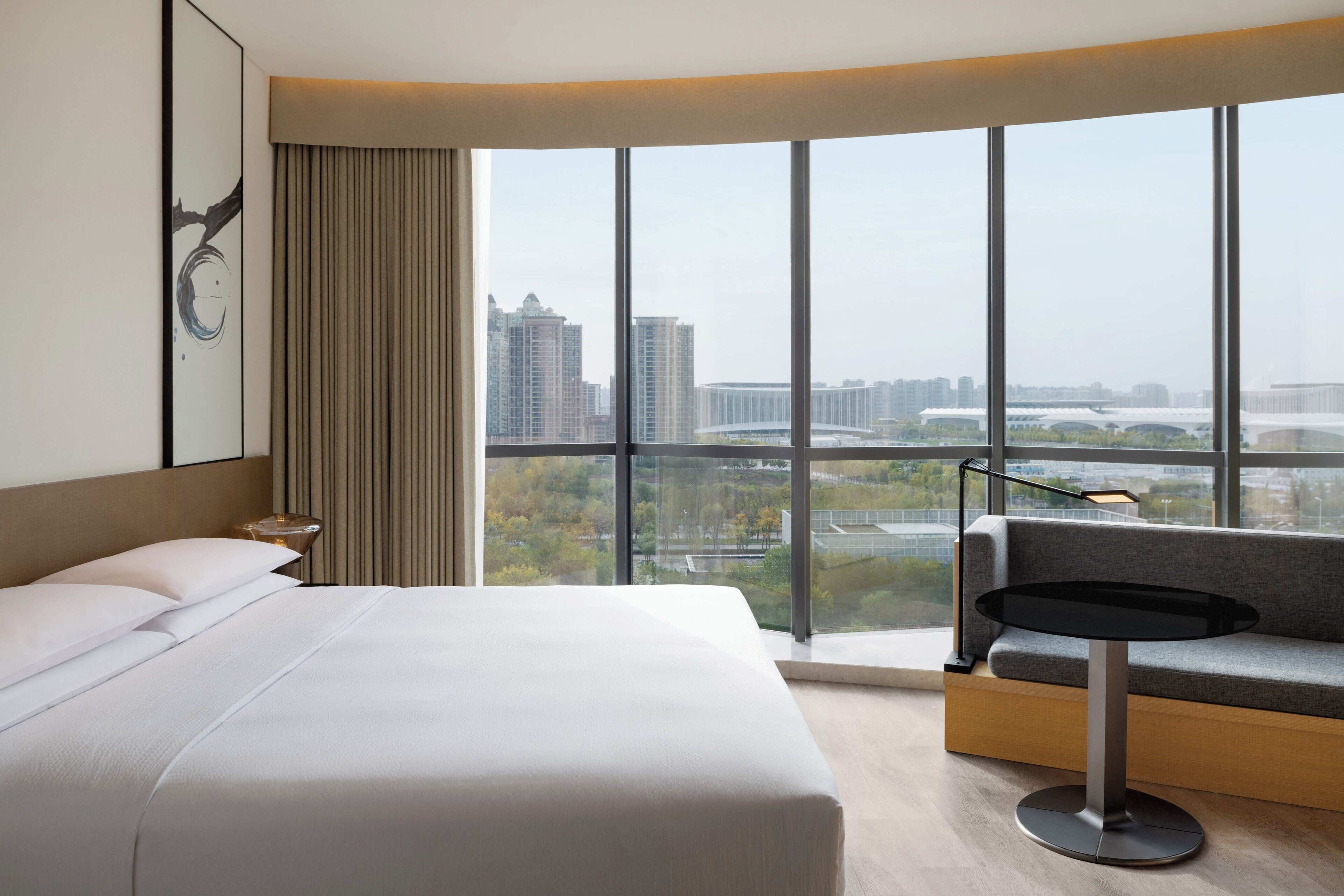A modern hotel room with a large bed, a sofa, a small table, and a view of the cityscape through large windows.