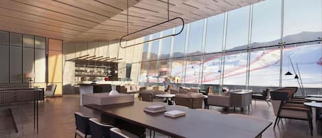 A spacious indoor area with a long table and chairs, a bar area, and large windows offering a view of a snowy landscape.