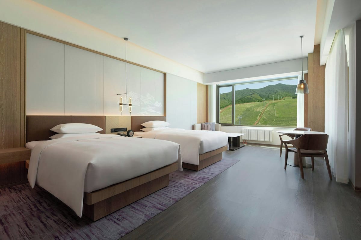 A modern hotel room with two beds, a dining area, and a large window offering a view of the countryside.