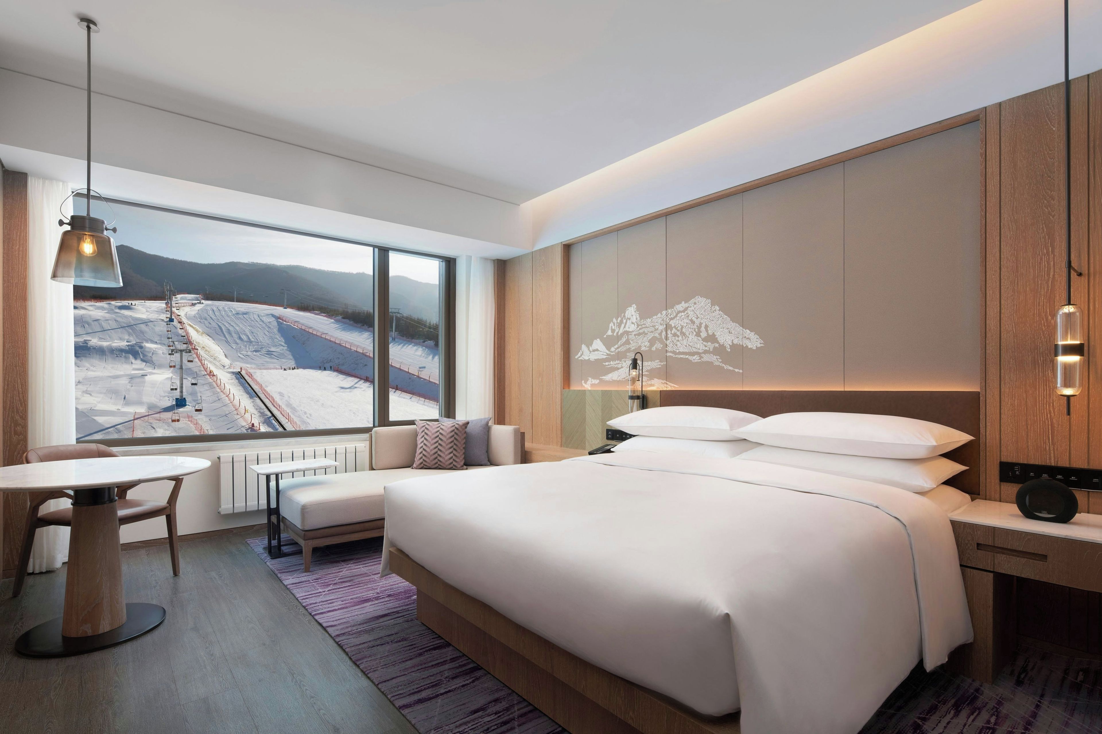 A modern hotel room with a large bed, a desk, a chair, and a view of a ski slope.