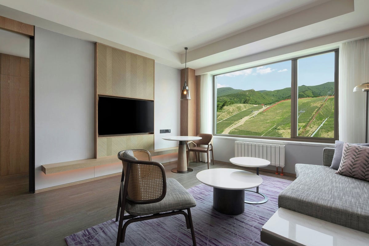 A modern room with a flat-screen TV, a sofa, a dining table, and a view of green hills.