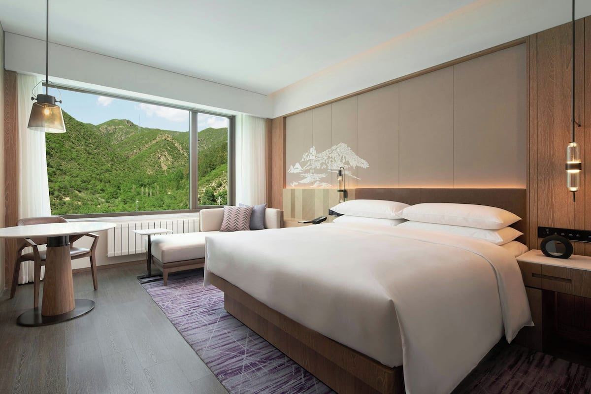 A modern hotel room with a large bed, a desk, a chair, a view of a mountainous landscape, and a large window.