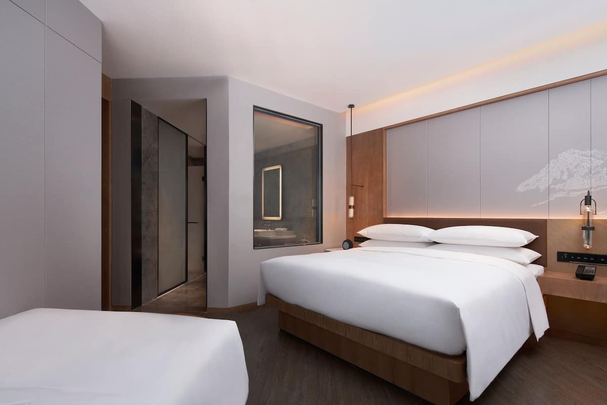 A modern hotel room with a large bed, a bedside table with a lamp, a mirror, and a view of a bathroom.