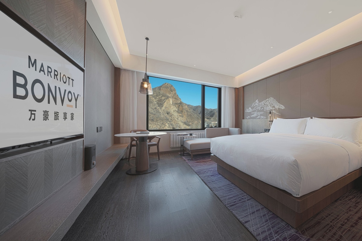 A modern hotel room with a large bed, a desk, and a view of the mountains.