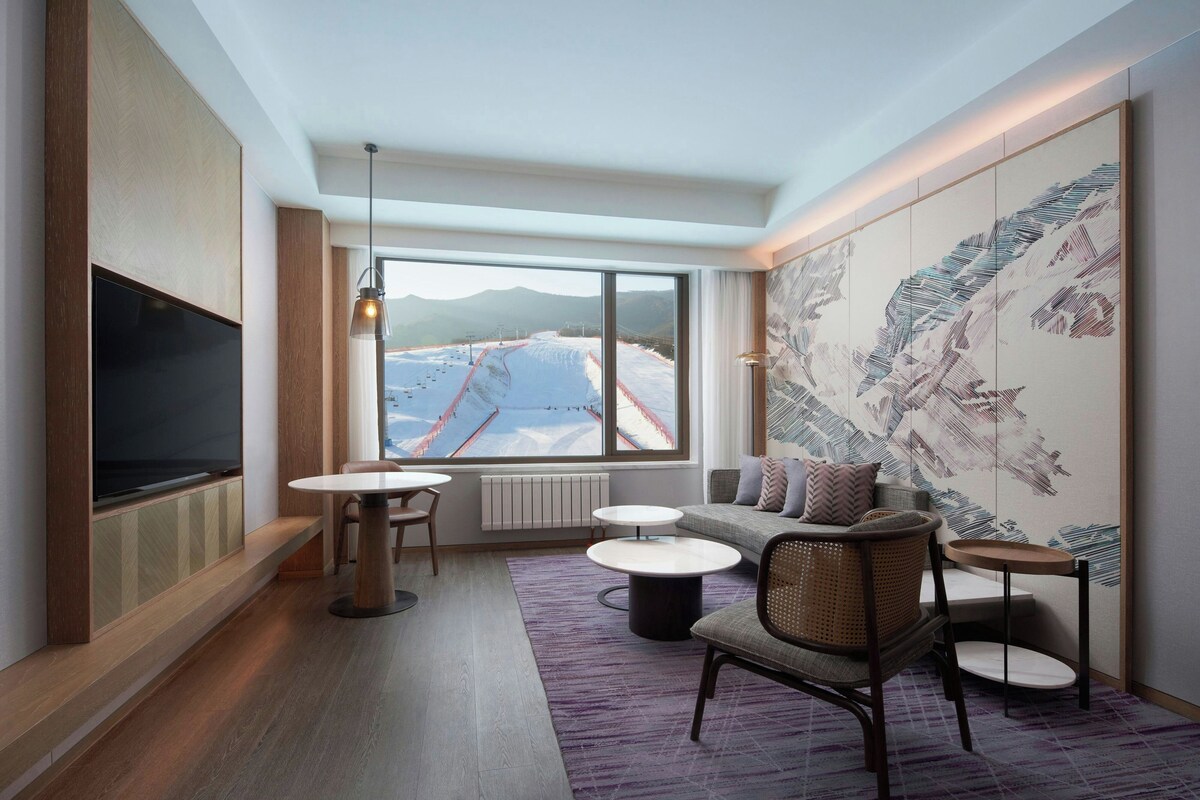A modern hotel room with a large window, a flat-screen TV, a sofa, a dining table, and a decorative wall.