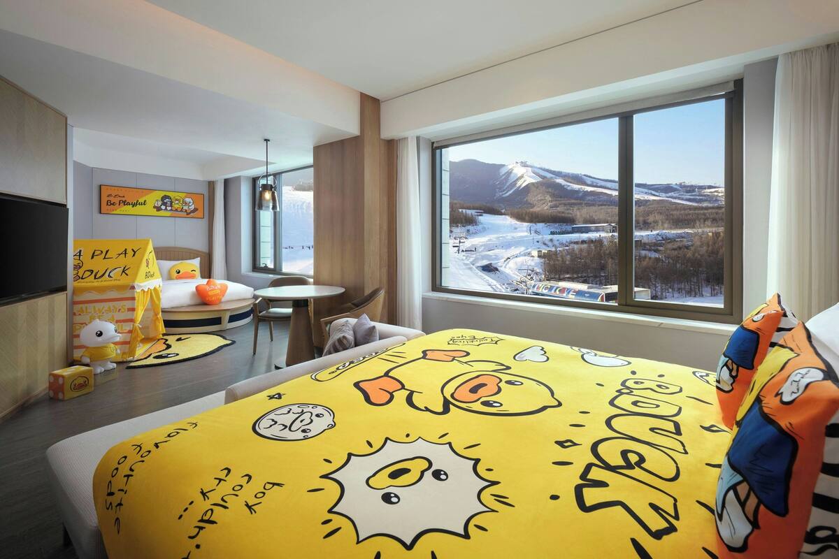 A hotel room with a large window offering a view of a snowy landscape, a bed with a yellow cartoon-themed bedspread, a small table with a lamp, and a TV on the wall.