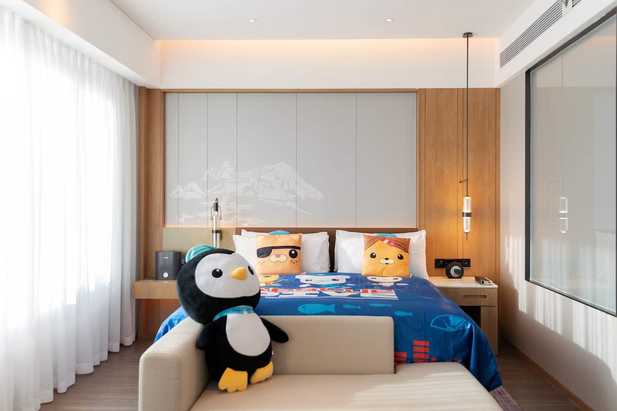 A modern hotel room with a large bed, a plush penguin toy, and a nightstand with a lamp.