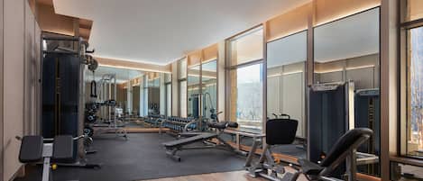 Un gimnasio moderno con diversos equipos de ejercicio, amplios espejos y vistas de la ciudad a través de grandes ventanales.