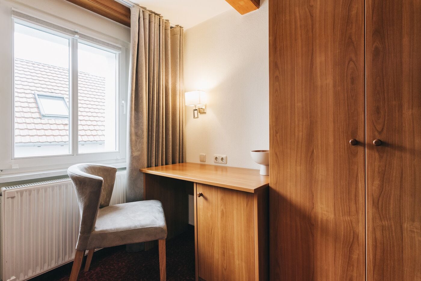 A hotel room with a wooden desk, a chair, a radiator, and a window with curtains.