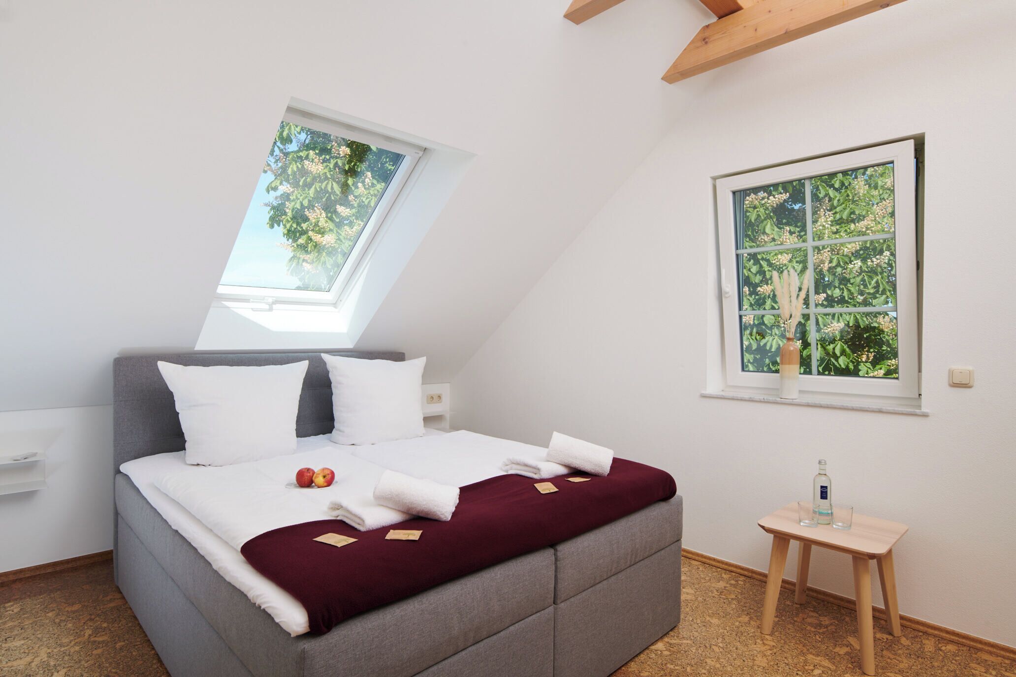 A bedroom with a bed, two windows, a small table, and a bottle of water.