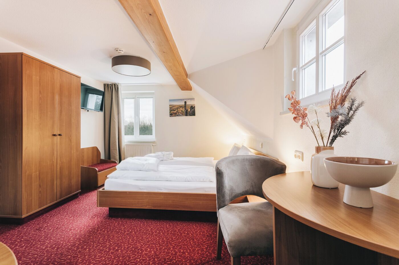 A hotel room with a bed, a wooden wardrobe, a desk with a bowl, a chair, and a window with a view of the outdoors.