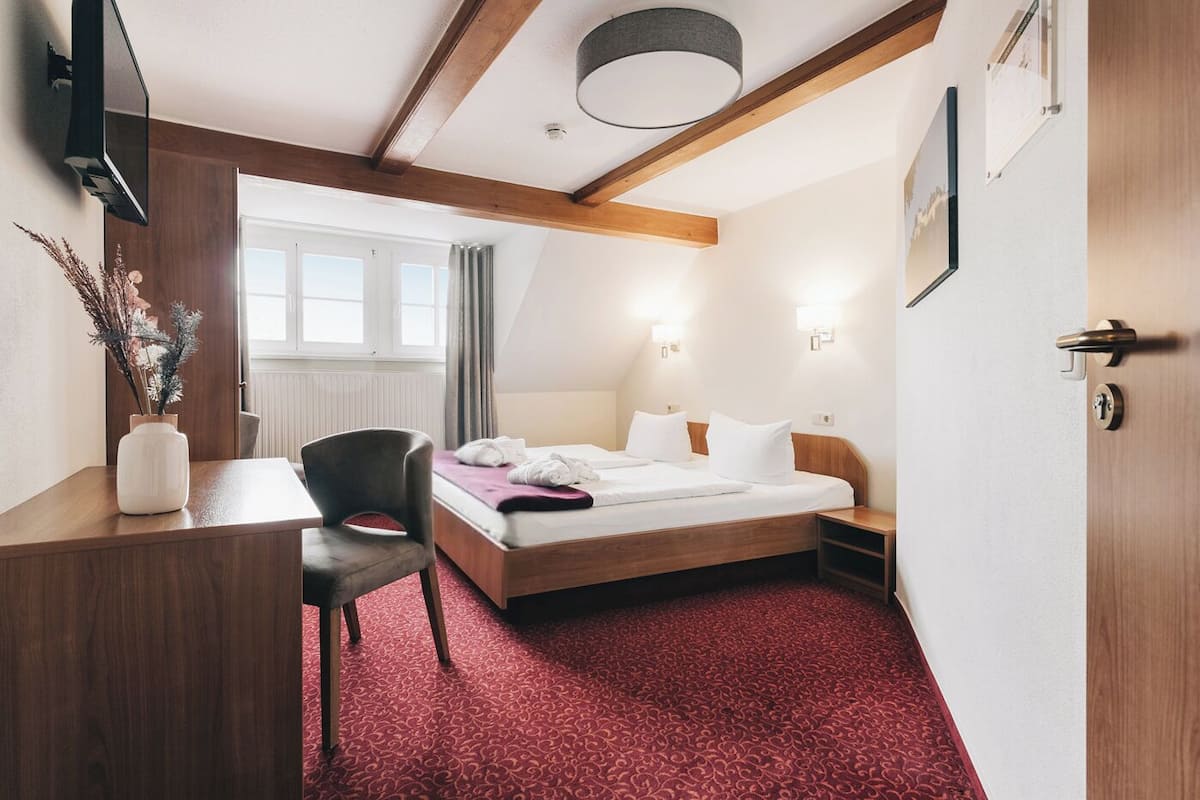 Une chambre d’hôtel avec un lit, un bureau, une chaise, un vase contenant des fleurs séchées et une fenêtre avec des rideaux.