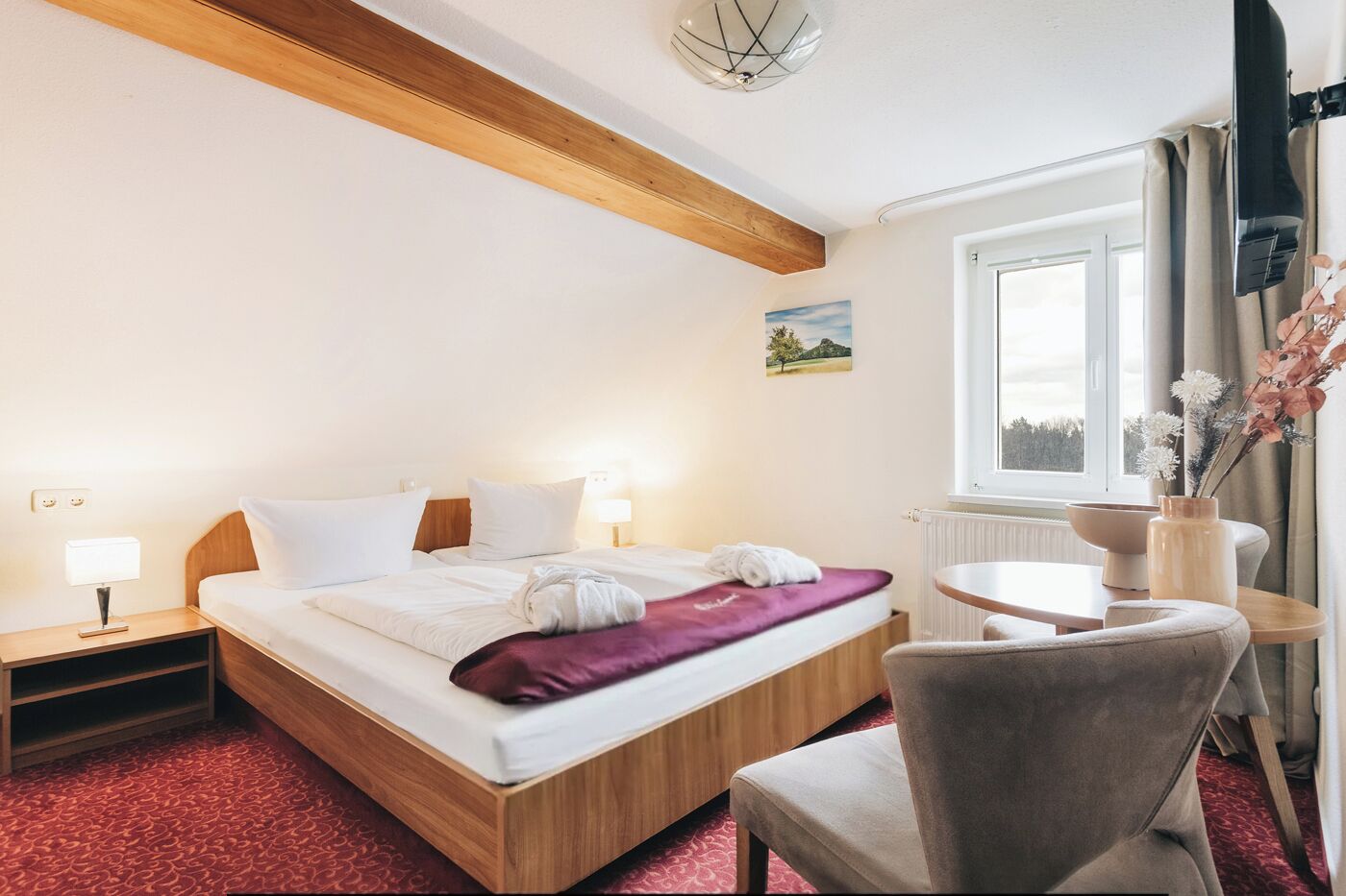 Habitación de hotel con una cama de madera, una mesita de noche con lámpara, una mesa redonda con un jarrón de flores y una ventana con cortinas.