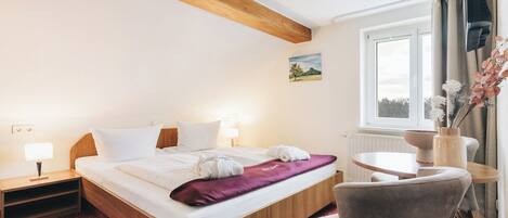 Habitación de hotel con una cama de madera, una mesita de noche con lámpara, una mesa redonda con un jarrón de flores y una ventana con cortinas.