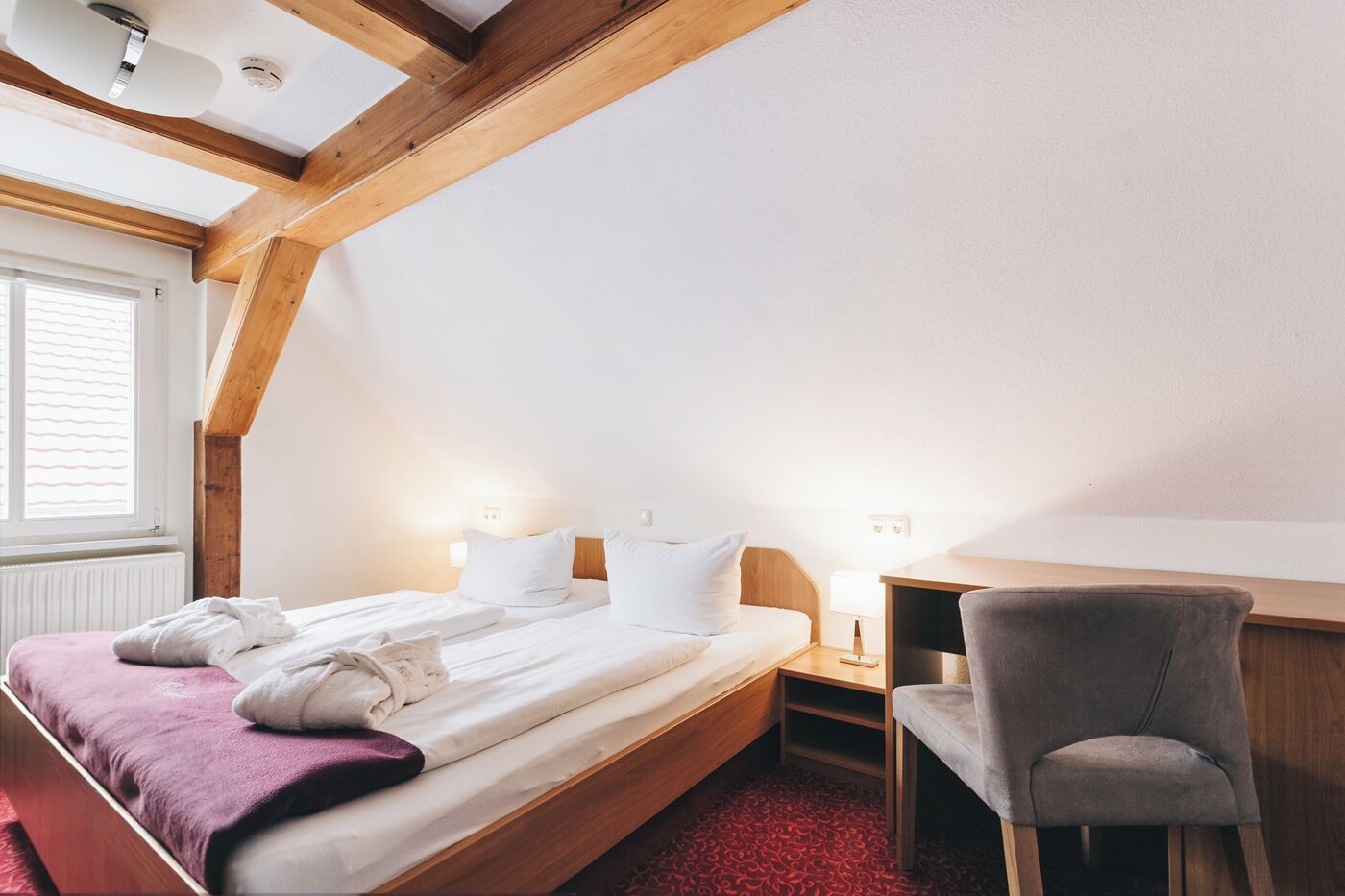 A hotel room with a wooden ceiling, a bed with white linens, a desk with a chair, and a window with blinds.