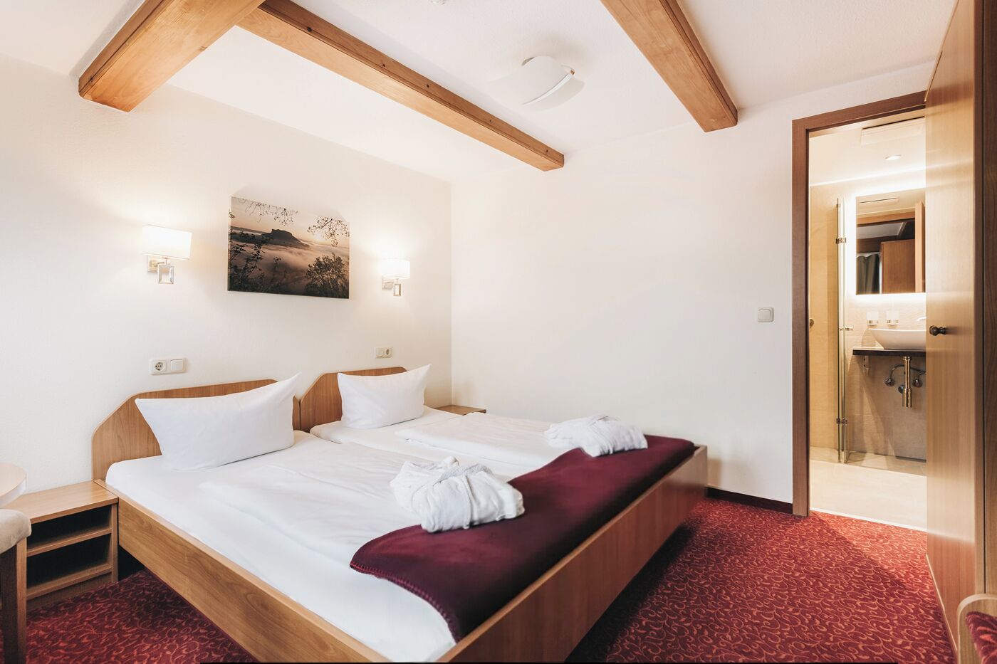 Habitación de hotel con una cama grande, cabecero de madera, dos mesitas de noche, alfombra roja, un cuadro en la pared y un baño visible a través de una puerta abierta.