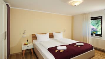 A double bed with white bedding and a maroon throw, a nightstand with a bottle and glasses, and a window with a view of greenery.