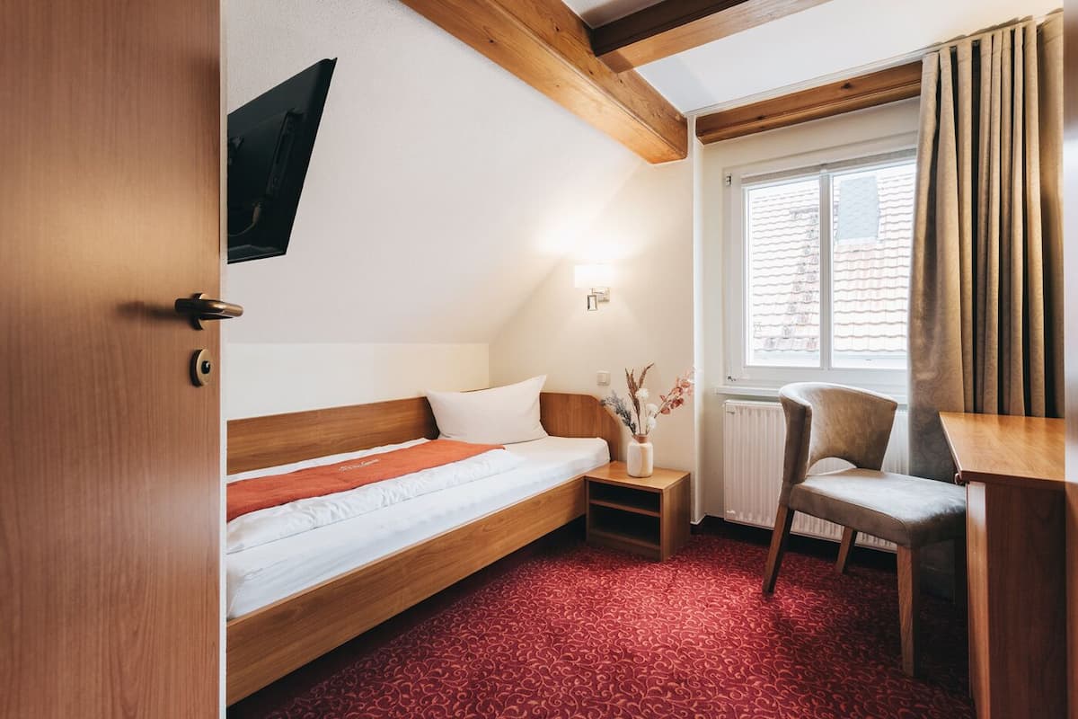 Une chambre d’hôtel avec un lit en bois, un bureau, une chaise, une fenêtre avec des rideaux et un radiateur.