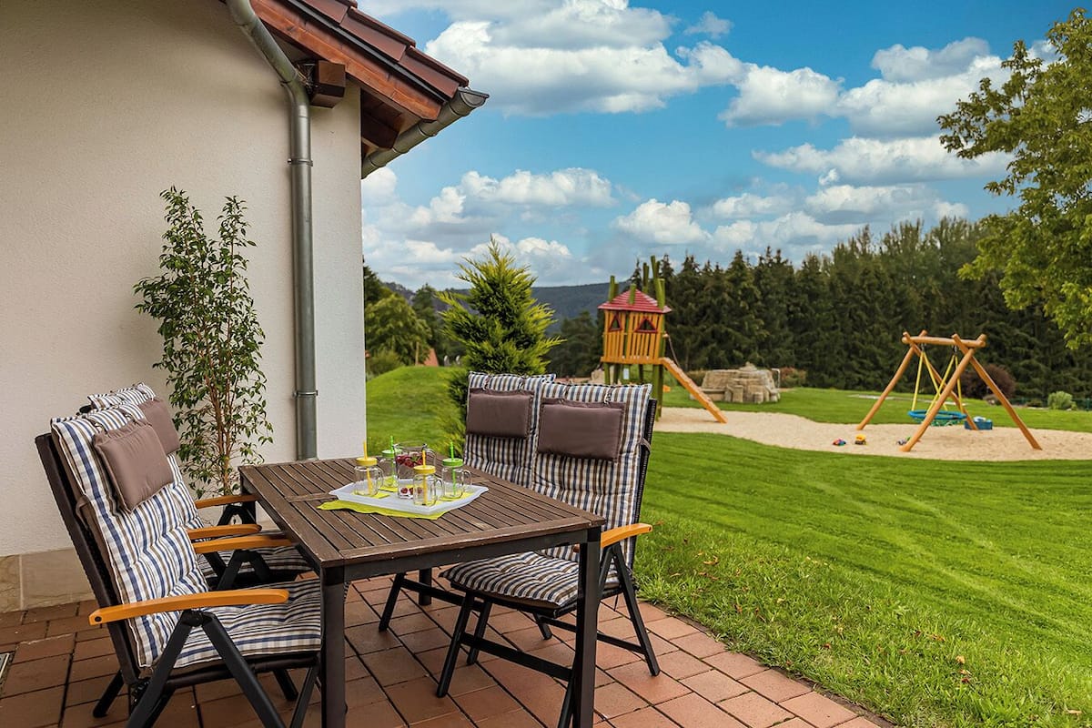 Une terrasse avec une table et des chaises, un espace de jeux avec une balançoire et une cabane, et une pelouse verdoyante.