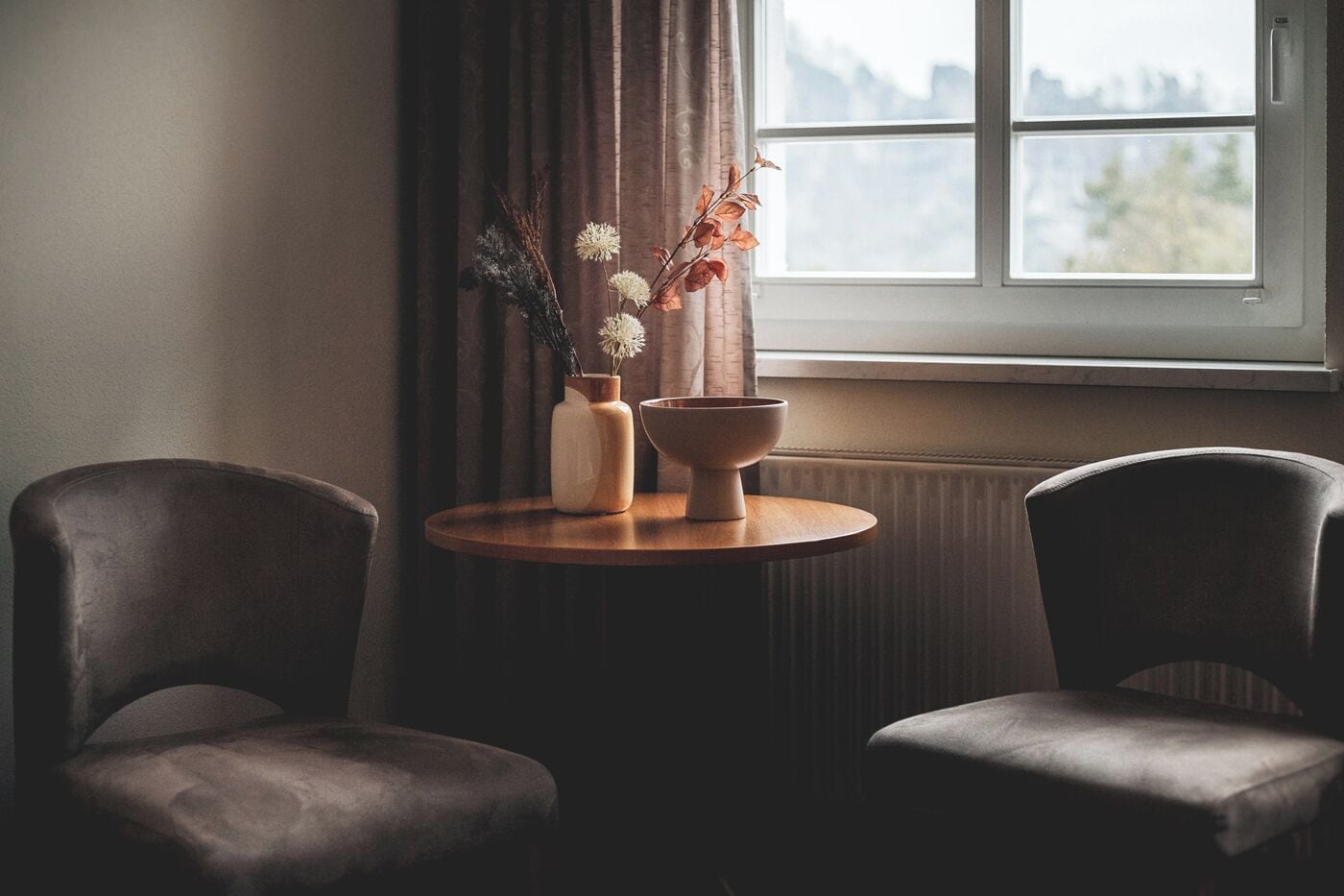A room with a round wooden table, two dark chairs, a vase with flowers, and a window with curtains.