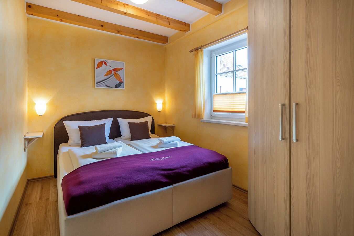 A bedroom with a bed, a window, a painting, and wooden beams on the ceiling.