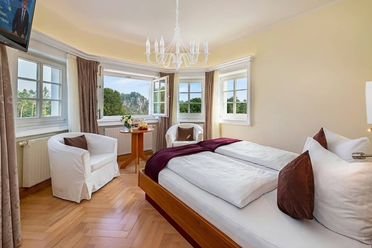 Une chambre avec un grand lit, un fauteuil blanc, une petite table et une vue sur l’extérieur.