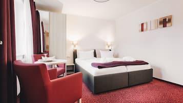 Una camera d'albergo con un letto, due comodini, una scrivania con una sedia, un tappeto rosso e uno specchio.
