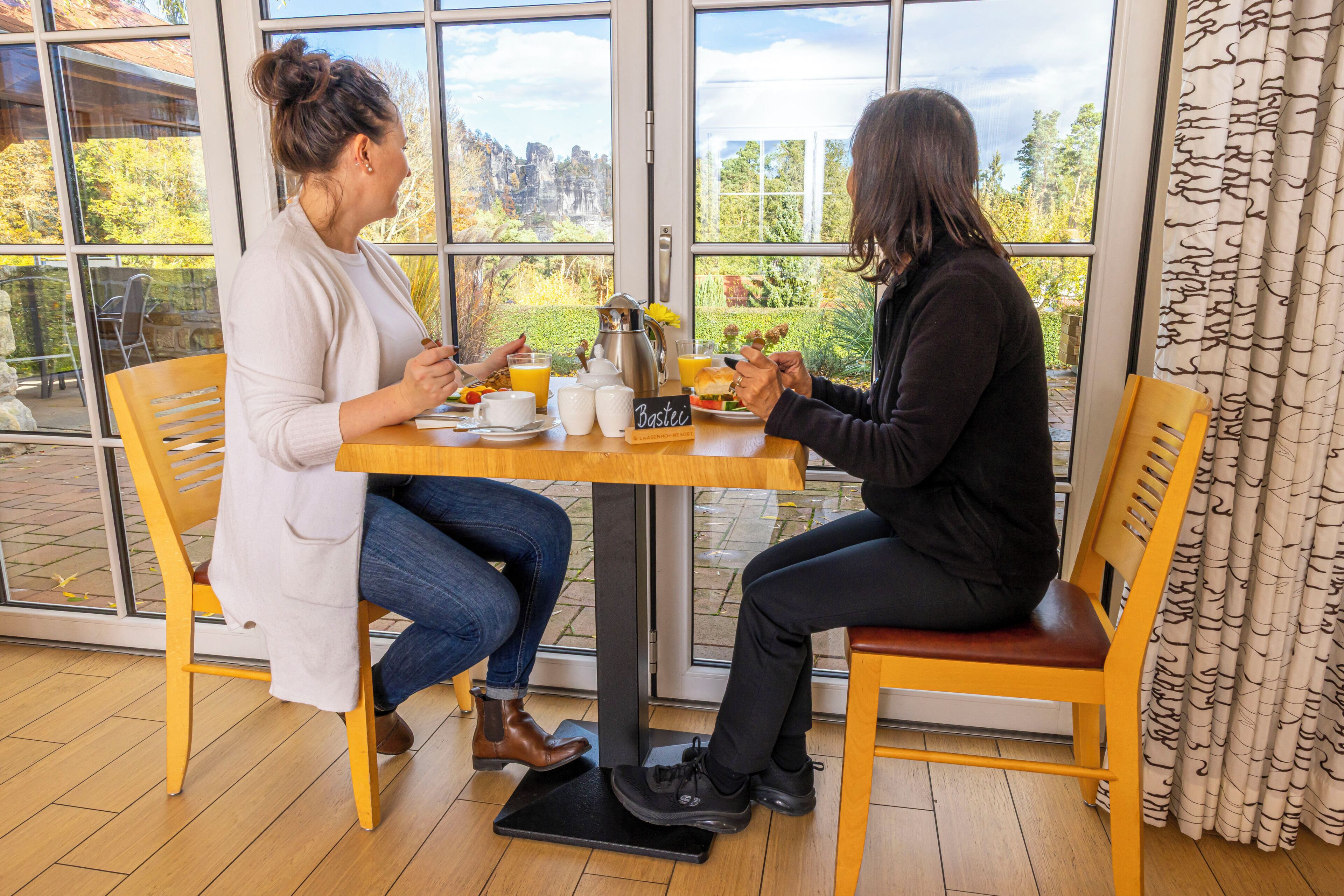 Dos personas sentadas en una mesa de madera, con vista a árboles y un edificio a través de amplias ventanas.