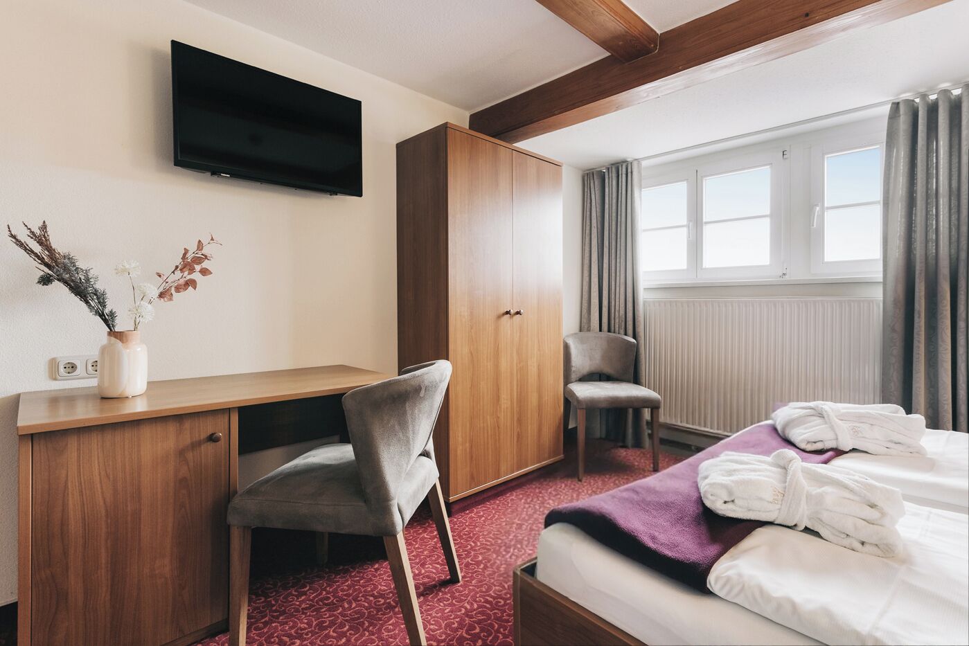 A hotel room with a bed, a desk, a chair, a wardrobe, a TV, and a window with curtains.