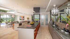 A modern kitchen with a large island, a dining area with a view, and a large window. - Villa De Claris, Brand New Villa (Uvita)