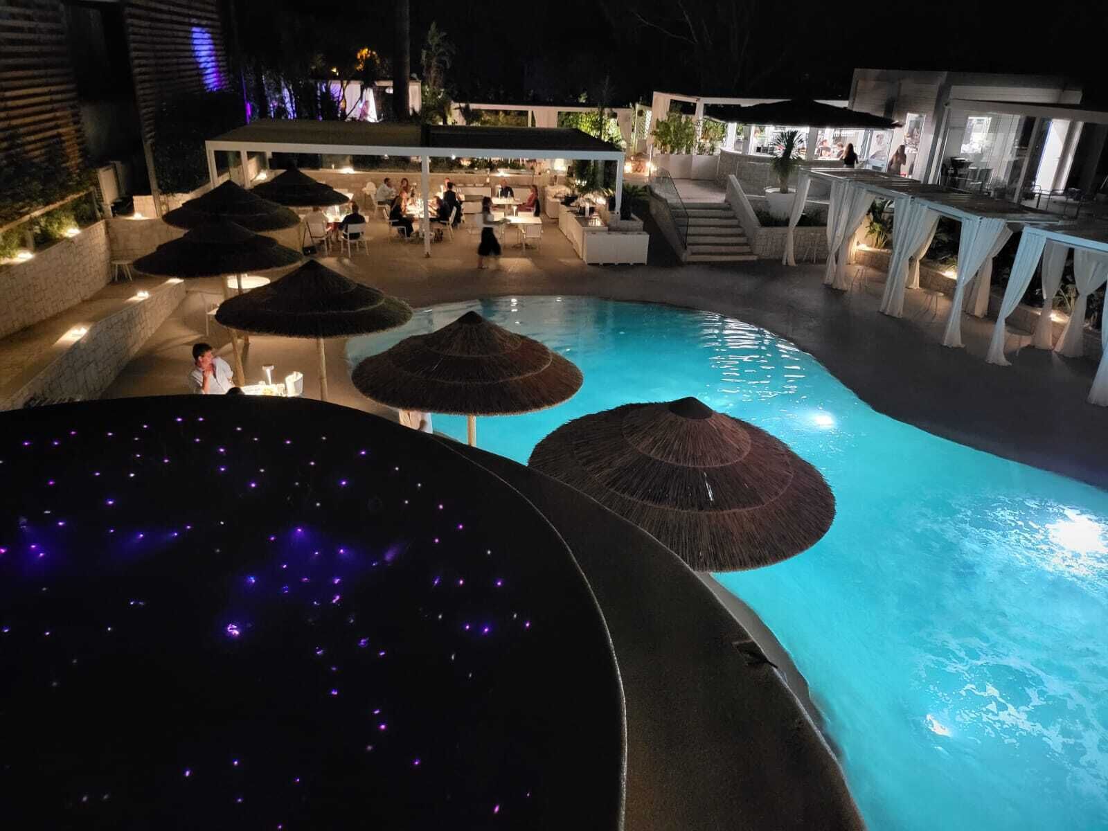 Zona de piscina nocturna con sombrillas y bañera de hidromasaje.